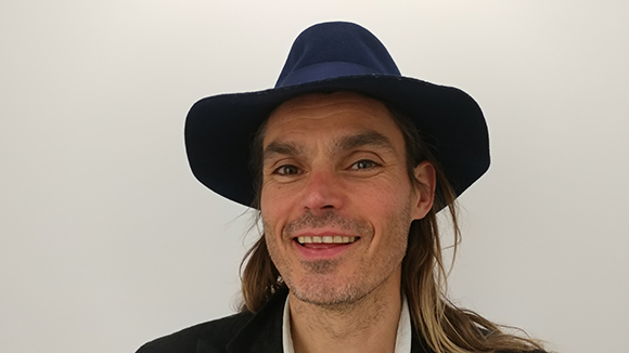 A smiling man with long hair wearing a dark blue wide-brimmed hat against a white background.