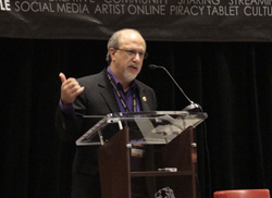 A man in a dark suit and purple shirt gestures while speaking into a microphone at a podium.