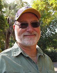 A man with a white beard, wearing a yellow and purple cap and sunglasses, smiling in front of green trees.