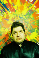 A man in a black jacket looks upward in front of a bright, multicolored abstract paint splatter background.