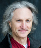 A man with long, wavy grey hair and blue eyes smiling slightly against a dark background.