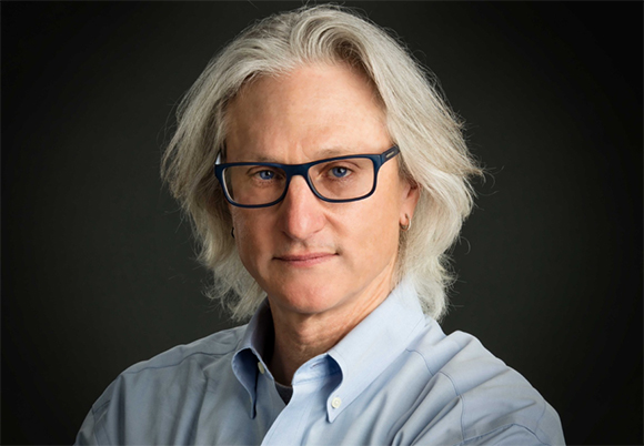 A man with long grey hair and blue-rimmed glasses wearing a light blue button-down shirt.