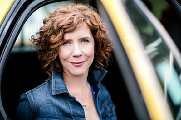 A woman with curly red hair and a denim jacket smiling while sitting in a vehicle.