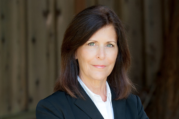 A woman with shoulder-length brown hair wearing a black blazer and white shirt looks toward the camera.