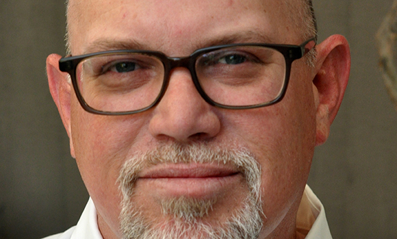Close-up of a man with short hair, dark-rimmed glasses, and a grey goatee looking toward the camera.