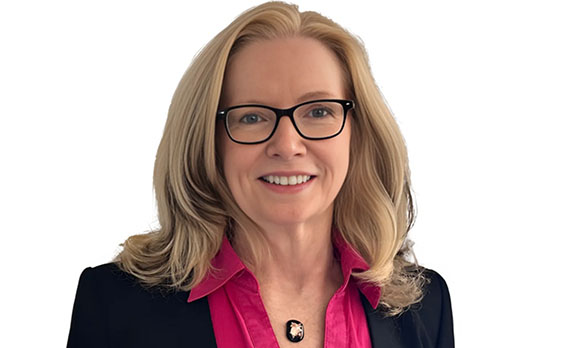 A smiling blonde woman with black-rimmed glasses wearing a pink collared shirt and black blazer.
