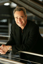 A smiling man with graying hair wearing a black blazer leans on a metal railing.