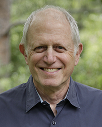 A smiling older man with thinning grey hair wearing a dark button-down shirt against a blurred green background.