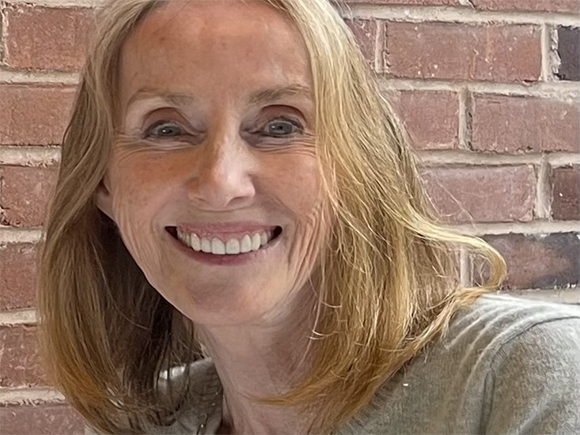 A smiling woman with shoulder-length blonde hair posing in front of a red brick wall.