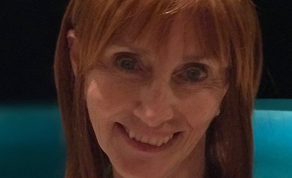A close-up portrait of a smiling woman with reddish-brown hair and bangs.