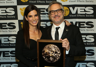 A woman in black and a man in a suit and glasses hold a gold award in front of a Visual Effects Society backdrop.