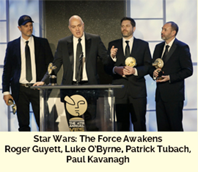 Four men in dark suits stand on a stage holding gold awards behind a podium.