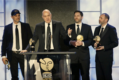 Four men in dark suits stand on a stage; one speaks at a podium while others hold gold awards.