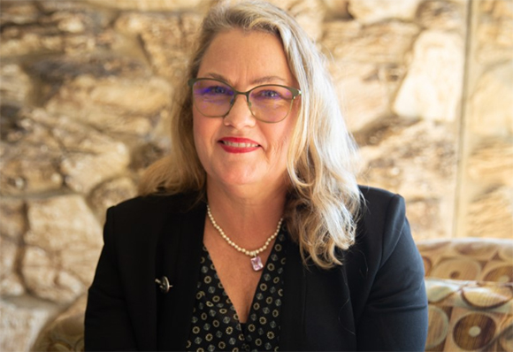 A blonde woman in a black blazer and pearl necklace smiles against a stone wall background.