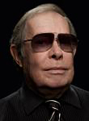 A man with light hair wearing dark tinted sunglasses and a black collared shirt.