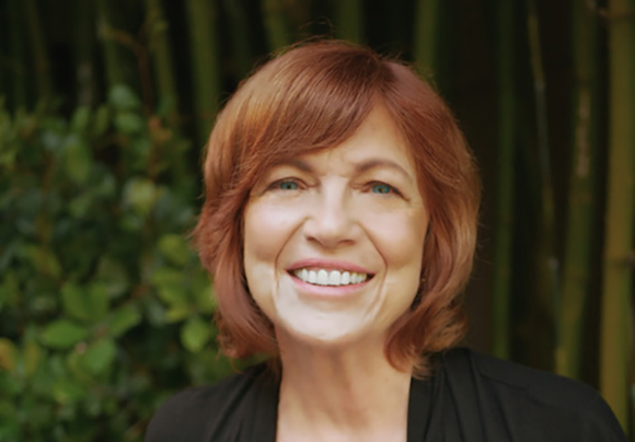 A smiling woman with shoulder-length reddish-brown hair and blue eyes, wearing a black top.