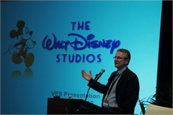 A man in a suit gestures while speaking at a podium in front of a large Walt Disney Studios screen.