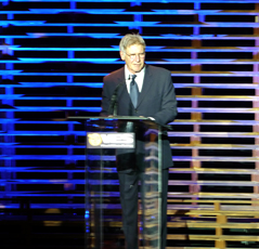 A man in a dark suit and tie stands behind a clear podium against a blue and gold striped background.