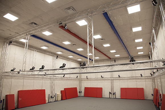 A large studio with high ceilings, metal trussing, overhead lights, and red mats along the walls.