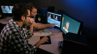 Two men sit at a desk with multiple computer monitors displaying video content in a dimly lit room.