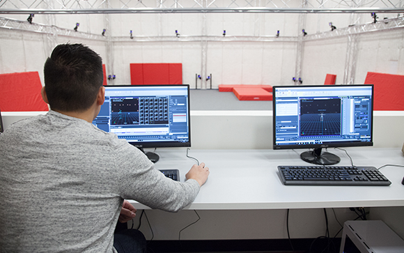 A person sits at a white desk with two computer monitors facing a large, empty studio with red mats.