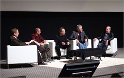 Five men sitting in white armchairs on a stage during a panel discussion against a black background.