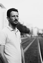 Black and white portrait of a man in a polo shirt standing outdoors near railway tracks.