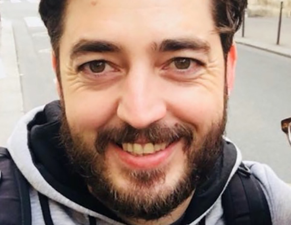Close-up of a smiling man with dark hair and a beard wearing a grey hoodie.