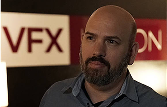 A bald man with a salt-and-pepper beard looking to the side in front of a sign that says VFX.