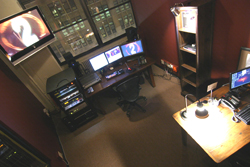 High-angle shot of a dark room containing multiple computer monitors, a desk with equipment, and a tall bookshelf.