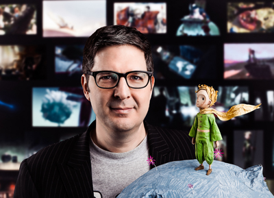 A man wearing black-rimmed glasses stands before a background of film frames next to a small animated figure.