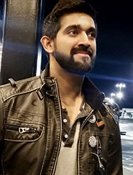 A man with dark hair and a beard wearing a brown leather jacket outdoors at night.