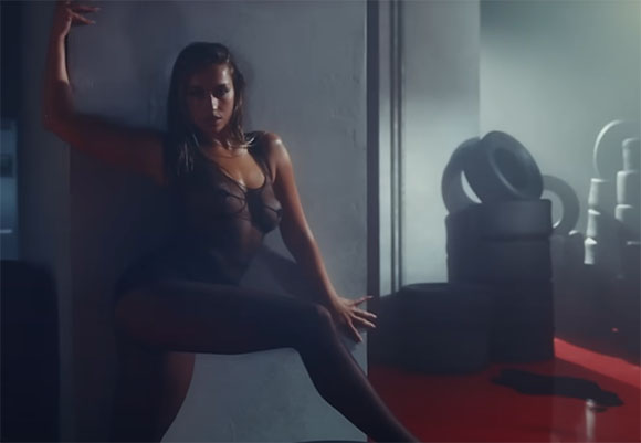 A woman in dark clothing poses against a wall near large tires in a dimly lit, smoky room.