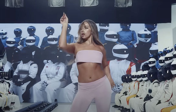 A woman in a pink crop top and leggings dances before a large backdrop of figures in white and blue helmets.