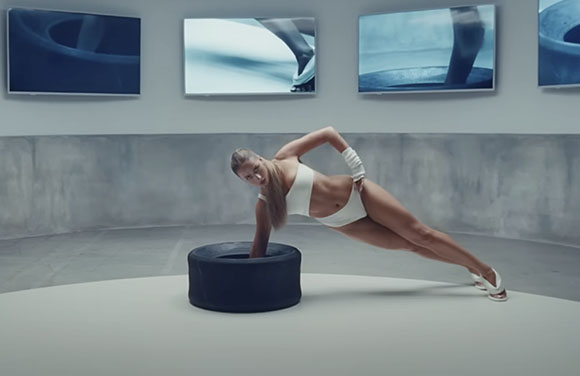 A woman in a white swimsuit holds a side plank pose with one hand on a black ring in a gray studio.