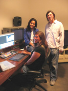 Three smiling people standing and sitting near a computer workstation with monitors and editing equipment.