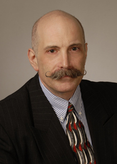 A bald man with a large, curled mustache wearing a dark pinstripe suit and patterned tie.