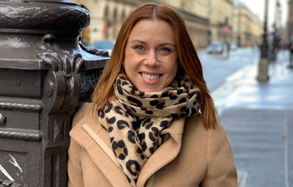 A smiling woman with reddish-brown hair wearing a tan coat and leopard print scarf outdoors.