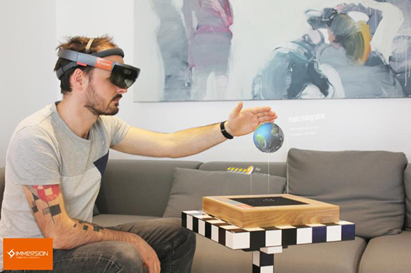 A man in a grey t-shirt uses a VR headset to interact with a floating holographic globe over a wooden base.