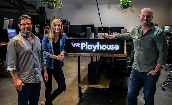 A man and woman stand on the left, while a man stands on the right of a glowing VR Playhouse sign.
