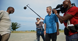 A camera operator and boom mic operator film a man in a blue shirt being interviewed outdoors.
