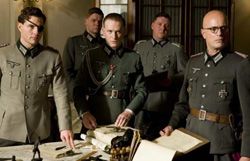 Five men in grey military uniforms stand around a table covered in papers and maps.