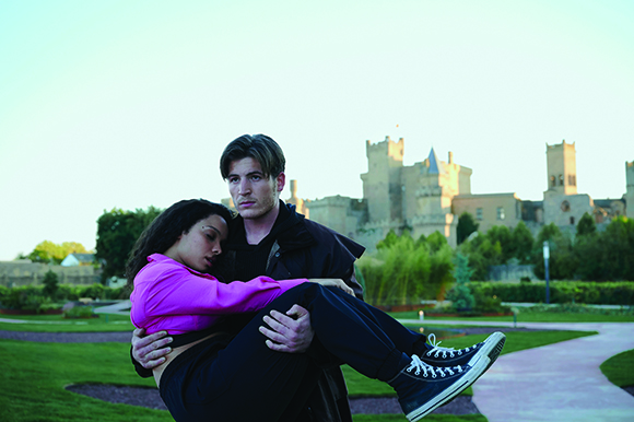 A man in a black jacket carries a woman in a pink shirt through a green garden toward a large castle.