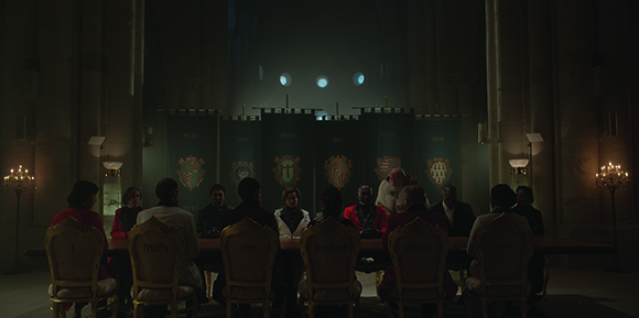 A group of people sit around a long table in a dimly lit, large hall with tall banners in the background.