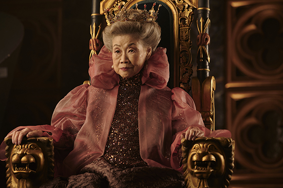 An elderly woman wearing a crown and pink gown sits on an ornate golden throne.