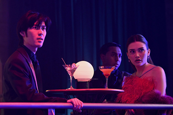 Three young people sit at a small table with cocktails under purple and blue lighting.