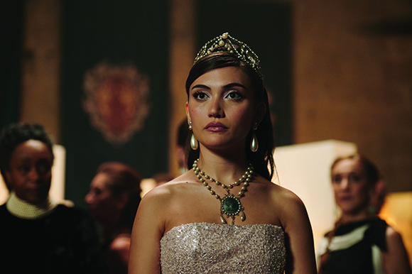 A young woman in a sparkling strapless gown, pearl necklace, and tiara looks ahead in a dimly lit room.