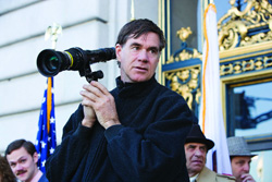 A man in a black sweater holds a large professional camera with a long lens in front of a building.