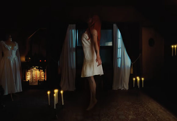 A person in a white dress levitates in a dark room lit by candles.