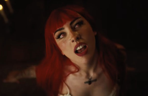 A woman with vibrant red hair, dark lipstick, and facial piercings looks upward with a dramatic expression.